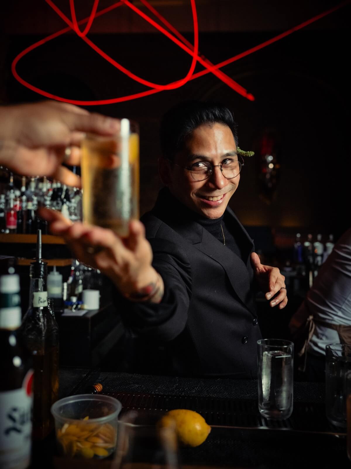 A man in round glasses offering a drink with red neon lights above him.