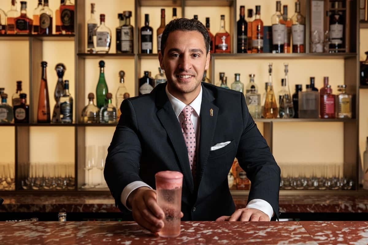 A bartender in a suit pushing a drink with a pink foam topping across the bar.