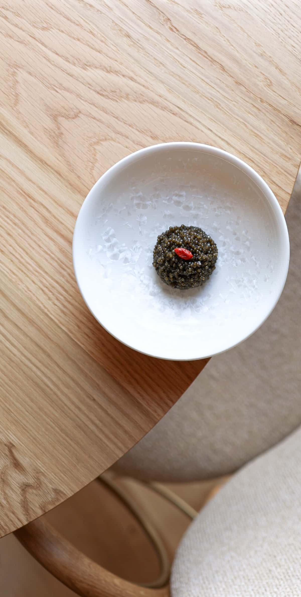 A wooden table, a white plate and a ball of caviar topped with a goji berry.